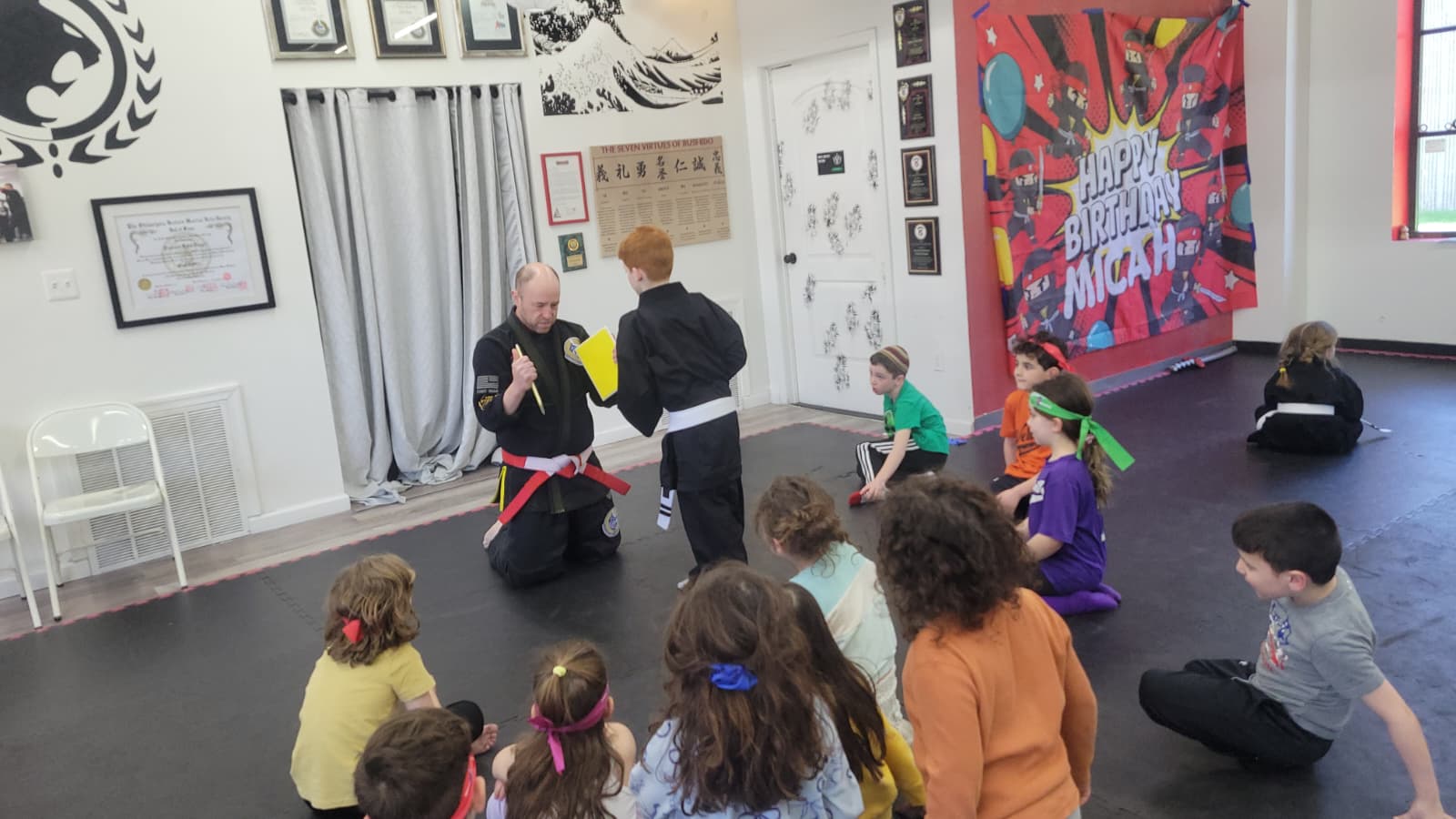 Instructor presenting a certificate to a child at a martial arts birthday party while other kids watch seated on the mat