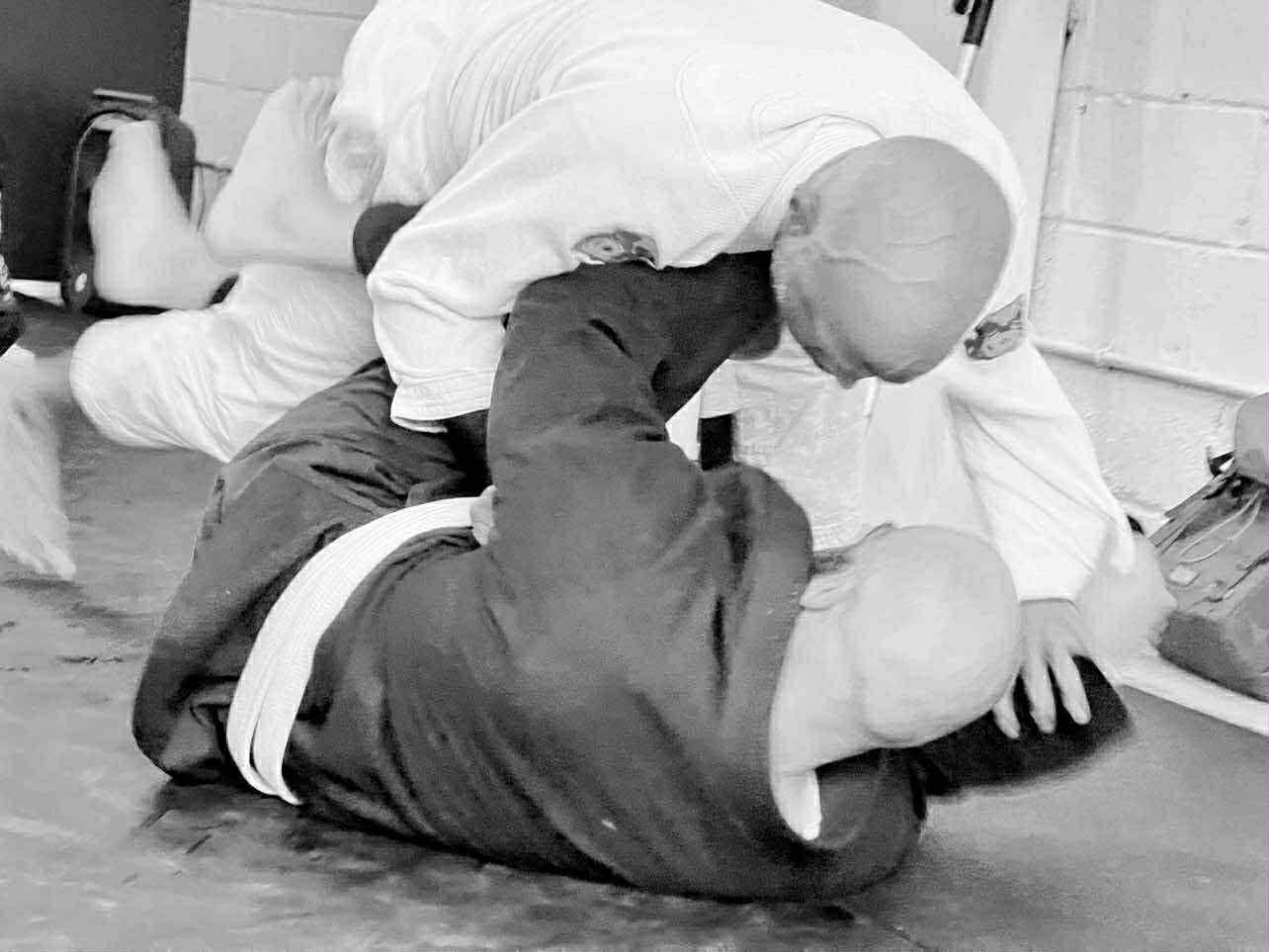 Two adult BJJ students in gis grappling in a top mount position on the mat, black and white photo